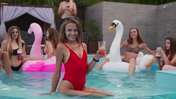 Mooie vrouw rode badpak drinken cocktail, opknoping uit met vrienden op 's nachts zwembad feest. Mensen die plezier hebben met dansen met drankjes en spetterend water in luxe privé villa zwembad. — Stockvideo