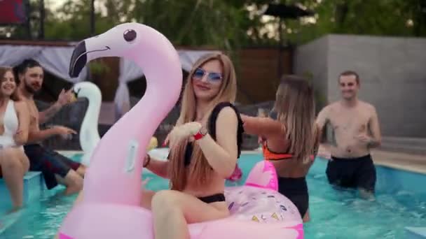 Mujer bonita en traje de baño de bikini colgando en el colchón de flamenco rosa inflable en la fiesta de la piscina de la noche. Amigos de fiesta con cócteles en villa de vacaciones. — Vídeo de stock