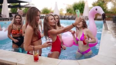 Girls in bikini have pool party with cocktails in swimming pool. Women relaxing clinking glasses with drinks at luxury resort. Female friends in red swimwear dancing, clubbing in a water. Slow motion.