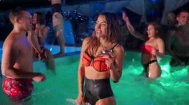 Bikinili güzel bir kadın kokteyl içiyor ve gece havuzu partisinde arkadaşlarıyla takılıyor. İnsanlar lüks özel villa havuzunda içkilerle dans edip su sıçratarak eğleniyorlar. Yavaş çekim.