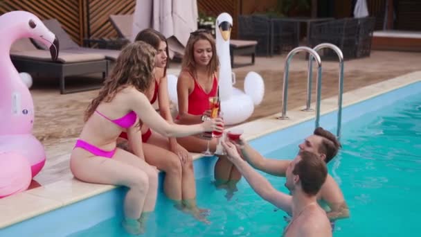 Attractive women in bikini clinking glasses with cocktails sitting by the swimming pool and flirting with fitted guys in a water. Girls with fresh colorful drinks relax on poolside in luxury resort. — Wideo stockowe