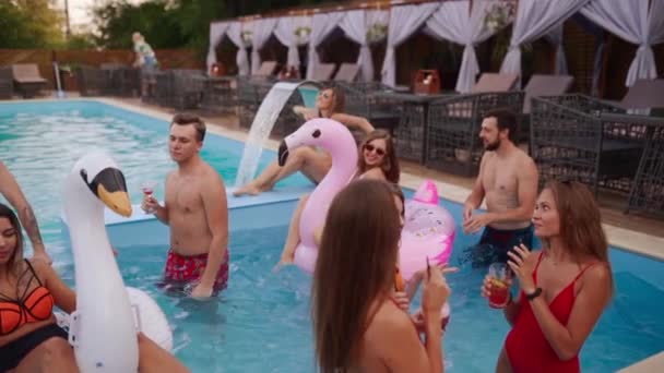 People have swimming pool party with cocktails at a luxury resort. Friends in swimwear enjoying drinks, hanging out and clubbing with inflatable floats. Hot girls relax in the water. Slow motion. — Video