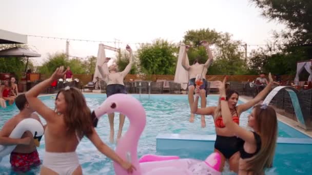 Друзья танцуют в частном бассейне виллы. Веселые молодые люди в купальниках прыгают и ныряют с брызгами воды в роскошном курорте. Мужчины и женщины тусуются в замедленной съемке. — стоковое видео