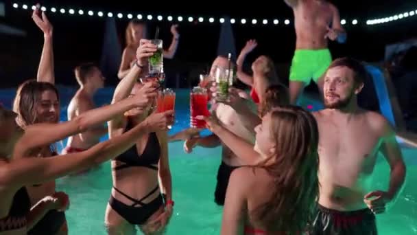 Group of friends toasting, clinking glasses with cocktails at night pool party. Cheerful people in swimwear drinking beverages, dancing, clubbing and partying in luxury resort villa. Slow motion. — Wideo stockowe