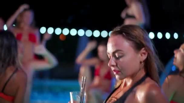 Hot woman in bikini drinking cocktail and hanging out with friends at night pool party. People having fun dancing with drinks and splashing water in luxury private villa swimming pool. Slow motion. — Vídeo de Stock