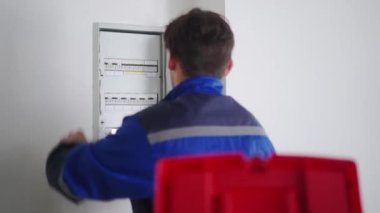 Elektrikçi elektrik sigorta kutusunu söker. Üniformalı usta elektriği tamir etmek için elektrik panelini erteliyor. Tamirci şalterleri ve şalterleri denetleyerek iç sorunları düzeltiyor..