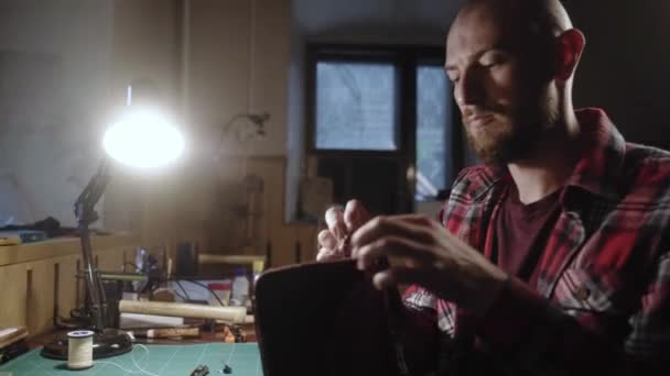 Skórzany rzemieślnik robi torebkę. Człowiek z igłą i nitką pracuje w warsztacie skórzanym. Krawiec w koszuli w kratę robi torebkę akcesoria do szycia w atelier. — Wideo stockowe