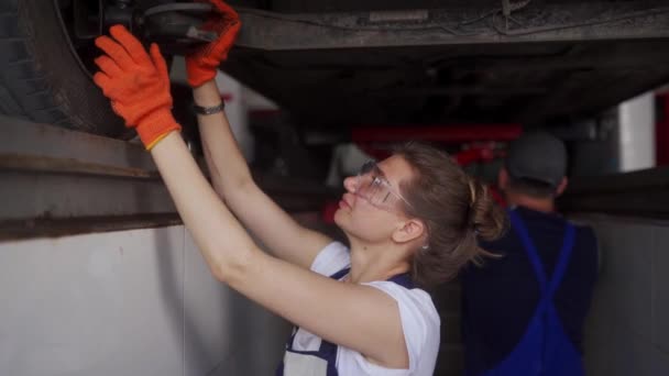 Samice mechanička kontroluje zavěšení auta. Silná žena trénuje mužského praktikanta. Pracovník kontroluje dno auta a podvozek vozidla na výtahu. Pracovníci v maskách kvůli karanténním omezením. — Stock video