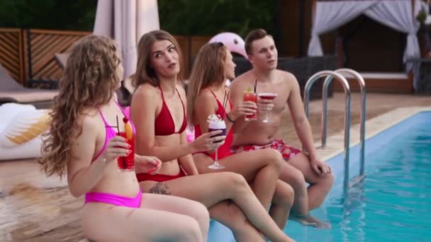 Atrakcyjne kobiety w bikini pijące świeże, kolorowe koktajle siedzące przy basenie i flirtujące z dopasowanym facetem w strojach kąpielowych. Dziewczyny relaksujące się na letniej imprezie przy basenie w luksusowym kurorcie. — Wideo stockowe