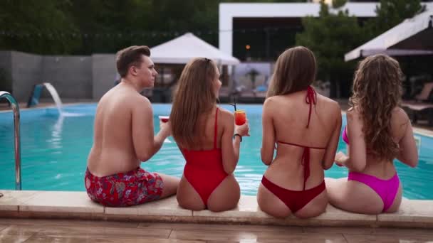 Attractive women in bikini drinking fresh colorful cocktails sitting by the swimming pool and flirting with fitted guy in swimwear. Girls relaxing on poolside summer party in luxury resort. — Stock Video