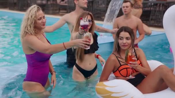 Ludzie urządzają imprezę przy basenie z koktajlami w luksusowym kurorcie. Przyjaciele w strojach kąpielowych, pijący drinki, spędzający czas z nadmuchiwanymi platformami. Gorące dziewczyny relaksują się w wodzie. Zwolniony ruch. — Wideo stockowe