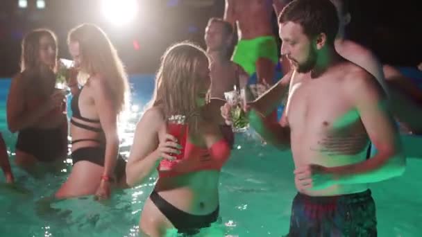 Group of friends toasting, clinking glasses with cocktails at night pool party. Cheerful people in swimwear drinking beverages, dancing, clubbing and partying in luxury resort villa. Slow motion. — Video