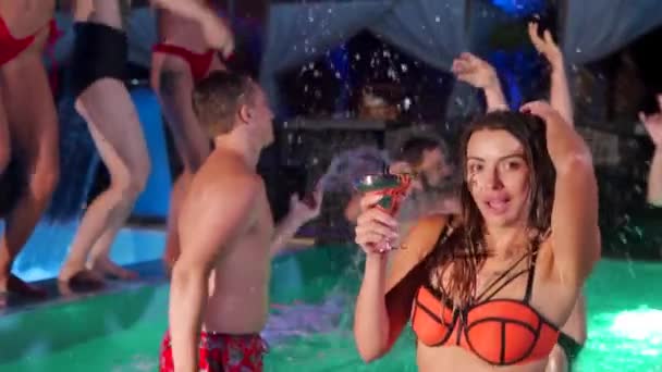 Красива жінка в бікіні п'є коктейль і висить з друзями на вечірці в басейні. Люди весело танцюють з напоями і розбризкують воду в розкішному приватному вілла басейні. Повільний рух . — стокове відео