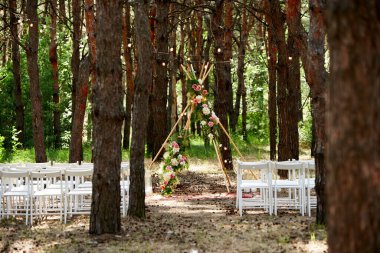 Çam ormanındaki açık hava düğün mekanında konili güzel bir bohem çadır kemeri süslemesi. Sandalyeler, çiçek kompozisyonları güller, halı, ip perisi ışıkları. Yaz kırsal düğünü.