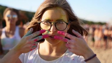Gözlüklü güzel bir kadın yanaklarına çizgiler çiziyor pembe renkli parmaklarıyla ağır çekimde. Kız Holi festivalinde eğleniyor. Kumsalda Hindu tatili. Koronavirüs karantinasının sonu