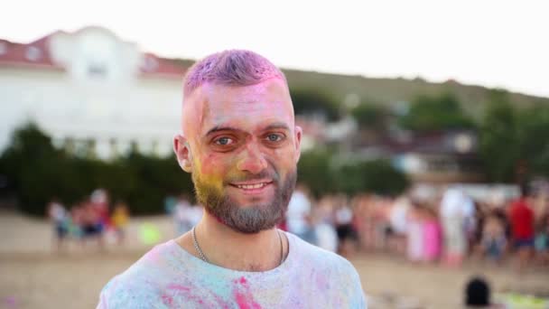 Smiling man with beard covered in colorful powder looks at camera. Cheeful guy smeared in dry colors at Holi festival. Outdoor hindu holiday party. End of lockdown, covid pandemic, restrictions. — Video