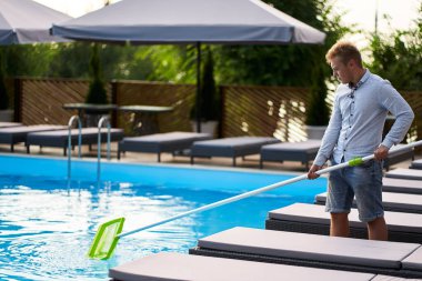 Çöpten yüzme havuzu temizleyen kaymak ağı ekipmanlı bir adam. Havuzun bakımı ve bakımı. Erkek, bir özsuyuyla çöpü sudan çıkarır. Yüzme havuzu temizleme servisi. Otel temizleyici.