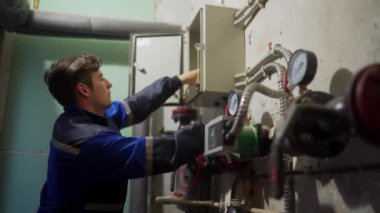 Elektrik denetleyicisi, bölgedeki ısıtma istasyonunun çoklu sayacıyla kontrol panelini inceliyor, uzaklaşıyor ve ışığı kapatıyor. Üniformalı usta teknik odada sigorta kutusunu test sondalarıyla kontrol ediyor..