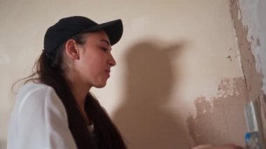 Profesyonel alçı dişisi duvarı alçılıyor, alçıyı tulum ve beyzbol şapkasıyla bir spatulaya sürüyor ve yayıyor. Duvarları sıva kaplı genç bir kadın, inşaat alanında dekorasyon işi yapıyor. İşlemler.