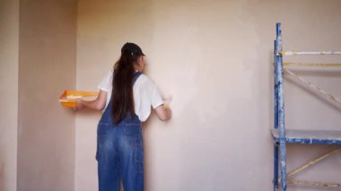 Profesyonel ressam kadın, inşaat alanında duvarı silindir ve tepsiyle boyuyor. Tulumlu ve şapkalı genç bir kadın ev dekorasyonu ve yenileme yapıyor. İşçi odada DIY onarımı yapıyor.