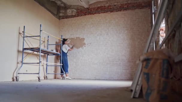 Professional plasterer female spackles the wall, applies and spreads plaster on a spatula in overalls and ball cap. Young woman plasters walls, doing decoration work on construction site. Occupations. — Stock Video