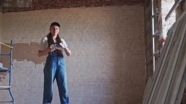 专业的灰泥匠女性会在墙壁上打孔，涂上石膏，并在工作服和球帽的水沫上撒布。年轻的女石膏墙壁，在建筑工地做装修工作。职业：. — 图库视频影像