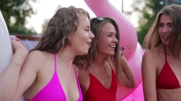 Heiße hübsche Mädchen in roter Badebekleidung spazieren mit aufblasbarem Flamingo, Schwan und Matratze am Pool. Attraktiv geschnittene Frauen im Bikini entspannen auf Sommerparty im Luxus-Resort. — Stockvideo