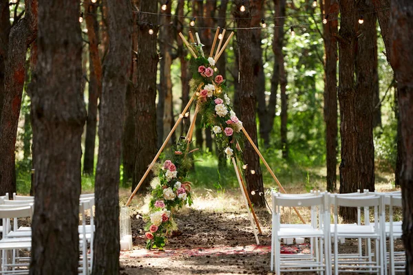Piękne cygańskie dekoracje łukowe tipi na zewnątrz miejsce ceremonii ślubnej w sosnowym lesie z szyszkami. Krzesła, kwiaciarskie kompozycje kwiatów róż, dywan, wróżkowe lampki smyczkowe. Letni ślub na wsi. — Zdjęcie stockowe