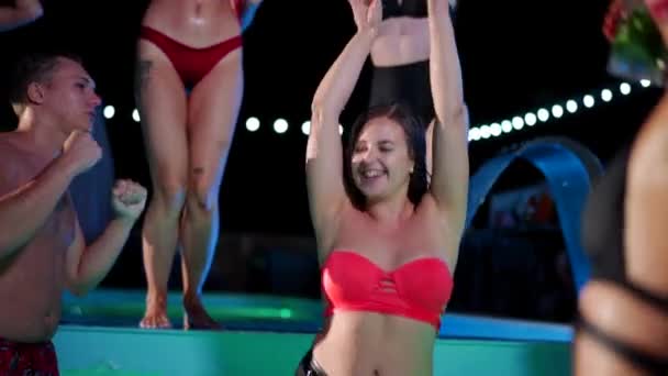 Hot woman in bikini dancing and hanging out with friends at night pool party. People having fun splashing water and partying with cocktails in luxury private villa swimming pool. Slow motion. — Stockvideo