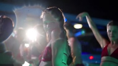 Hot woman in bikini dancing and hanging out with friends at night pool party. People having fun splashing water and partying with cocktails in luxury private villa swimming pool. Slow motion.