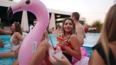 Hot girl in bikini hanging out with friends on pool party. Woman drink cocktail on inflatable pink flamingo float mattress on pool party. People partying and dancing in swimwear on tropical vacation.