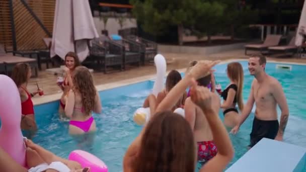 Mensen hebben een zwembadfeest met cocktails in een luxe resort. Vrienden in zwemkleding genieten van drankjes, rondhangen en uitgaan met opblaasbare praalwagens. Hete meisjes ontspannen in het water. Langzame beweging. — Stockvideo