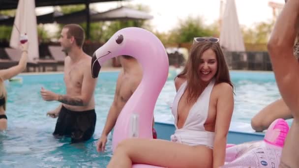 Гаряча дівчина в бікіні висить з друзями на вечірці біля басейну. Жінка розважається на надувному рожевому плаваючому матраці фламінго на вечірці в басейні. Люди вечірки і танці в купальнику на тропічній відпустці . — стокове відео