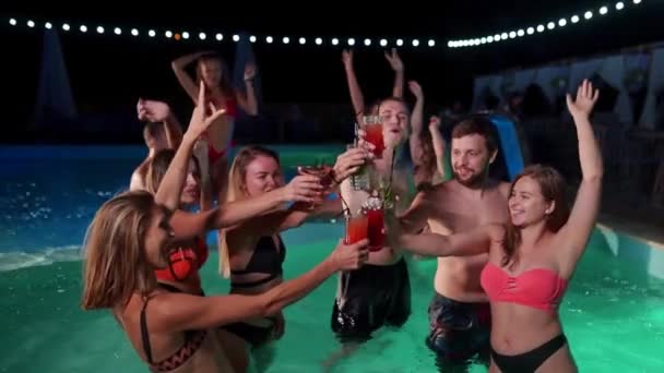 Group of friends toasting, clinking glasses with cocktails at night pool party. Cheerful people in swimwear drinking beverages, dancing, clubbing and partying in luxury resort villa. Slow motion. — Video Stock