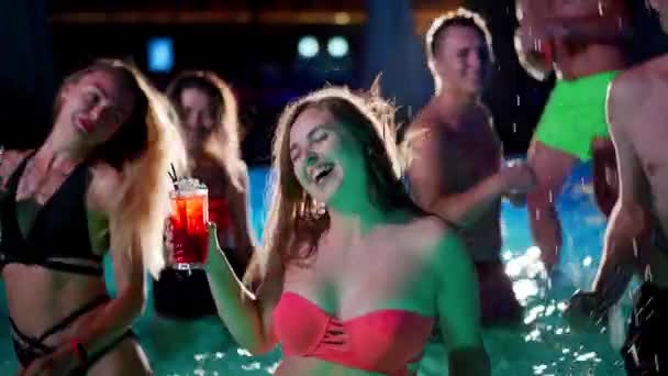 Pretty woman in bikini drinking cocktail and hanging out with friends at night pool party. People having fun dancing with drinks and splashing water in luxury private villa swimming pool. Slow motion. — Video Stock