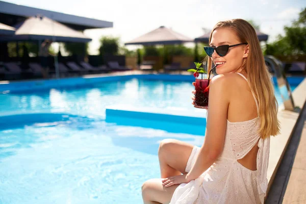 Blonde woman drinks refreshing cocktail sunbathing and sitting near swimming pool at tropical spa. Female in sunglasses enjoys a drink on poolside. Girl chilling in tropical resort on vacation. — ストック写真