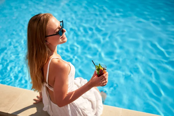Блондинка п'є освіжаючий коктейль засмагає і сидить біля басейну в тропічному спа-центрі. Жінка в сонцезахисних окулярах насолоджується випивкою на березі басейну. Дівчина охолоджує в тропічному курорті у відпустці . — стокове фото