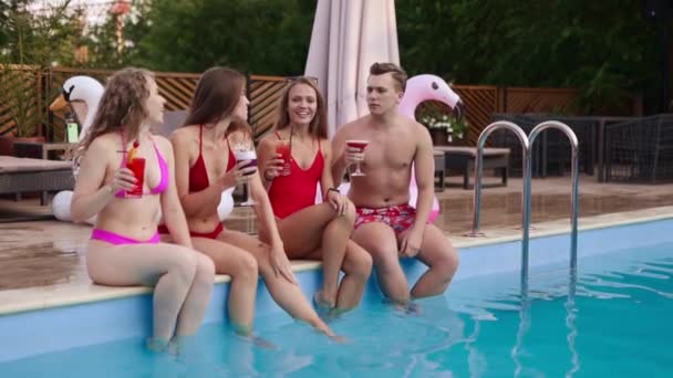 Atractivas mujeres en bikini tintineo vasos con cócteles sentados junto a la piscina y coqueteando con el chico equipado en traje de baño. Chicas con bebidas frescas y coloridas relajarse en la piscina en el complejo de lujo. — Vídeos de Stock