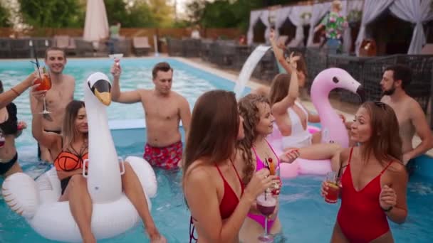Mensen hebben een zwembadfeest met cocktails in een luxe resort. Vrienden in zwemkleding genieten van drankjes, rondhangen en uitgaan met opblaasbare praalwagens. Hete meisjes ontspannen in het water. Langzame beweging. — Stockvideo