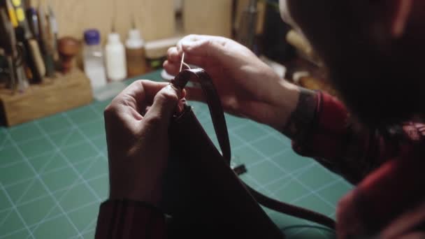 Läderhantverkare gör handväska. Man med nål och tråd arbetar i läderverkstad. Skräddarsydd skjorta i rutig skjorta vilket gör en väska sytillbehör i ateljén. Bakifrån. — Stockvideo