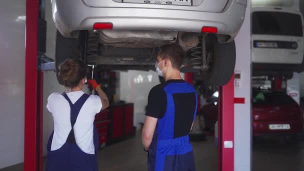 Mecánica femenina reparando la suspensión del coche. Mujer fuerte entrenar a un aprendiz masculino y trabajar con llave de enchufe con vehículo en un ascensor. Trabajadores con máscaras debido a restricciones de cuarentena. — Vídeo de stock