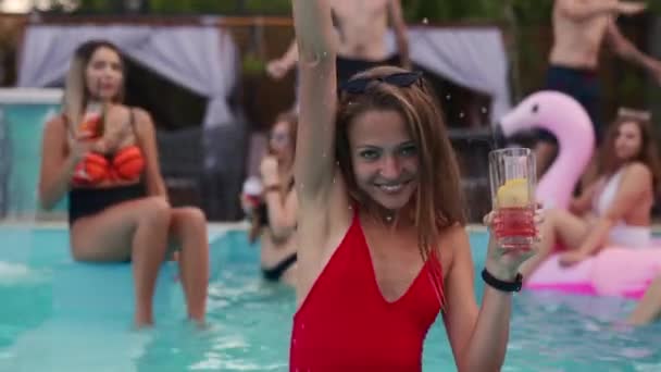 Mooie vrouw rode badpak drinken cocktail, opknoping uit met vrienden op 's nachts zwembad feest. Mensen die plezier hebben met dansen met drankjes en spetterend water in luxe privé villa zwembad. — Stockvideo