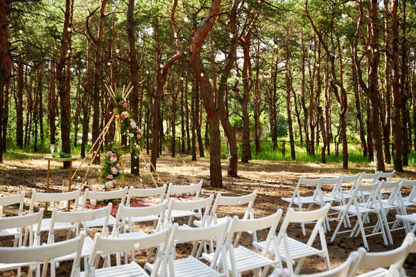 Çam ormanındaki açık hava düğün mekanında konili güzel bir bohem çadır kemeri süslemesi. Sandalyeler, çiçek kompozisyonları güller, halı, ip perisi ışıkları. Yaz kırsal düğünü.