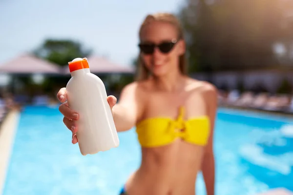 Happy woman shows suntan or sunscreen cream white bottle over the blurred swimming pool background. Tanned slim girl wearing yellow bikini swimwear and holds sun lotion. White bottle with copy space. — Stock Fotó