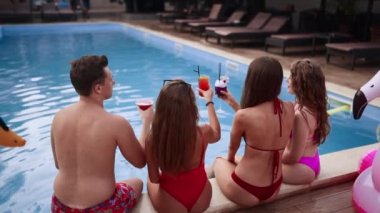 Bikini giyen, bardaklarını tokuşturup havuz kenarında oturan ve mayo giyen adamla flört eden çekici kadınlar. Taze renkli içkileri olan kızlar havuz başında dinlenirler. Arka plan.