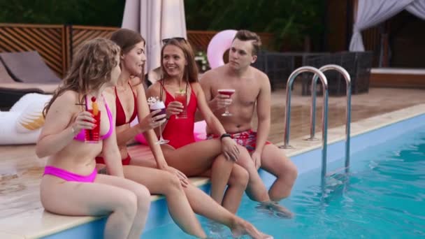 Attractive women in bikini drinking fresh colorful cocktails sitting by the swimming pool and flirting with fitted guy in swimwear. Girls relaxing on poolside summer party in luxury resort. — Stockvideo