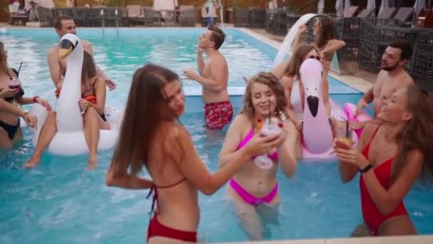 Die Leute feiern Poolparty mit Cocktails in einem Luxus-Resort. Freunde in Badebekleidung genießen Drinks, hängen herum und tummeln sich mit aufblasbaren Schwimmern. Heiße Mädchen entspannen im Wasser. Zeitlupe. — Stockvideo