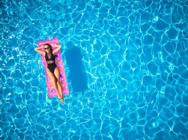Yüzme havuzunda bikinili güzel bir kız şişme pembe yatakta dinleniyor. Mayo bronzlaştırıcı giymiş zayıf seksi bir kadın. Lüks oteldeki mavi suda yüzen bir kadın. Gökyüzü, yukarıdan görünüm.