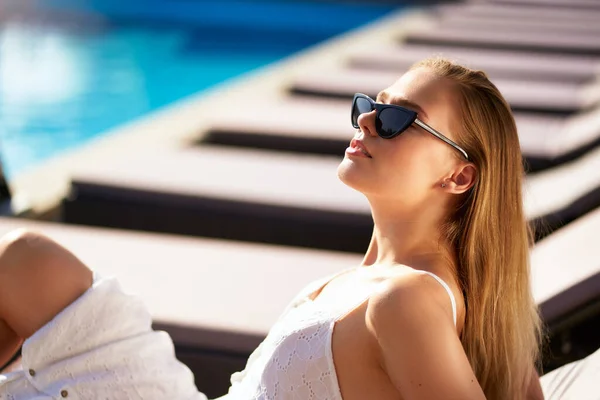 Красива жінка купається і охолоджується біля басейну на пляжному ліжку на тропічному курорті. Приваблива дівчина в білій сукні і сонцезахисних окулярах лежить на сонці на стільці на палубі в спа, розслабляючись в спекотний літній день . — стокове фото