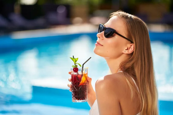 Mulher loira bebe refrescante cocktail banhos de sol e sentado perto da piscina no spa tropical. Feminino em óculos de sol gosta de uma bebida na beira da piscina. Menina refrigeração em resort tropical em férias. — Fotografia de Stock